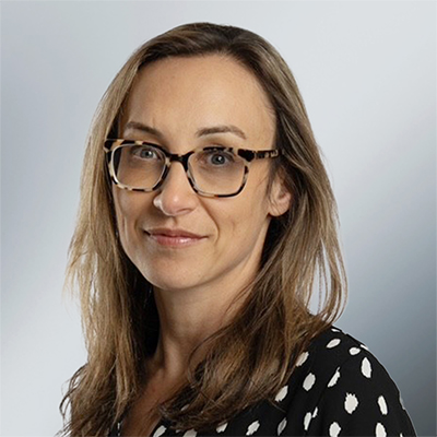 Image of Sara Gottlieb souris sur un fond gris, portant des lunettes et un habit professionnel