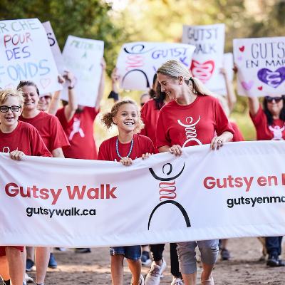 People in Gutsy Walk t-shirts carrying a Gutsy Walk sign. Words written on the image say, "Together as One. We walk to stop Crohn's and colitis. June 5th, 2022. gutsywalk.ca."